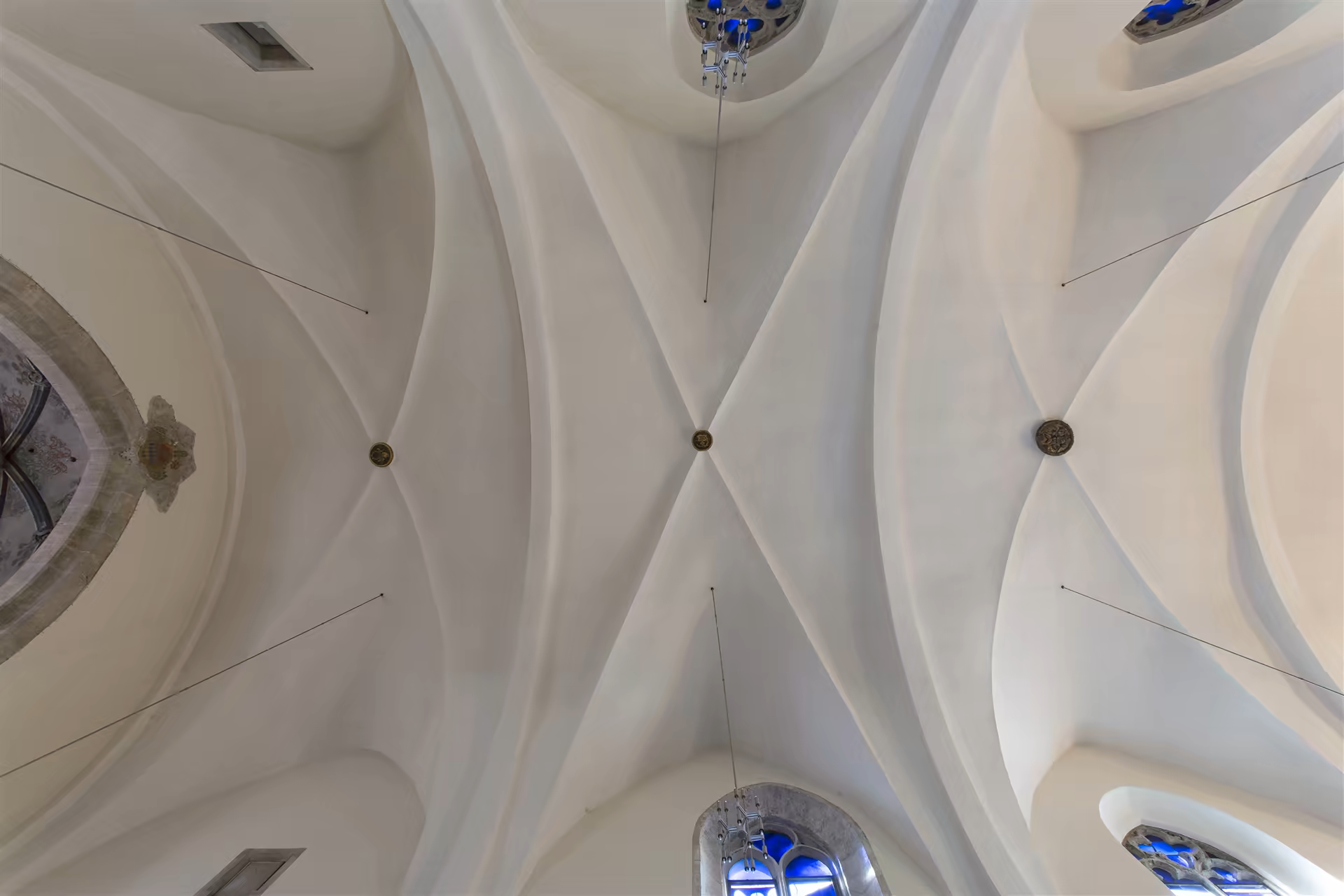 Plafond voûté en croisée d'ogives blanches avec clefs de voûte ornées dans une église, fenêtres en arc avec vitraux bleus partiels.