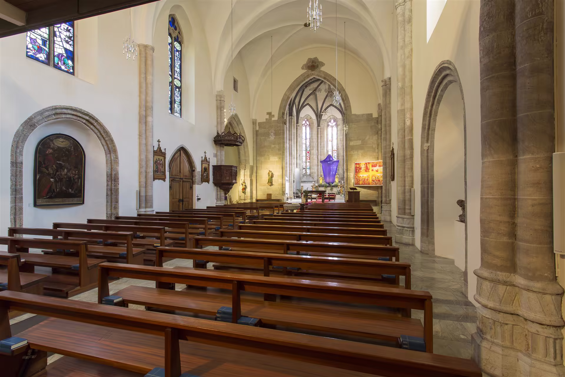 Intérieur d'une église avec rangées de bancs en bois, vitraux colorés, voûtes en pierre et autel orné d'un tissu violet.