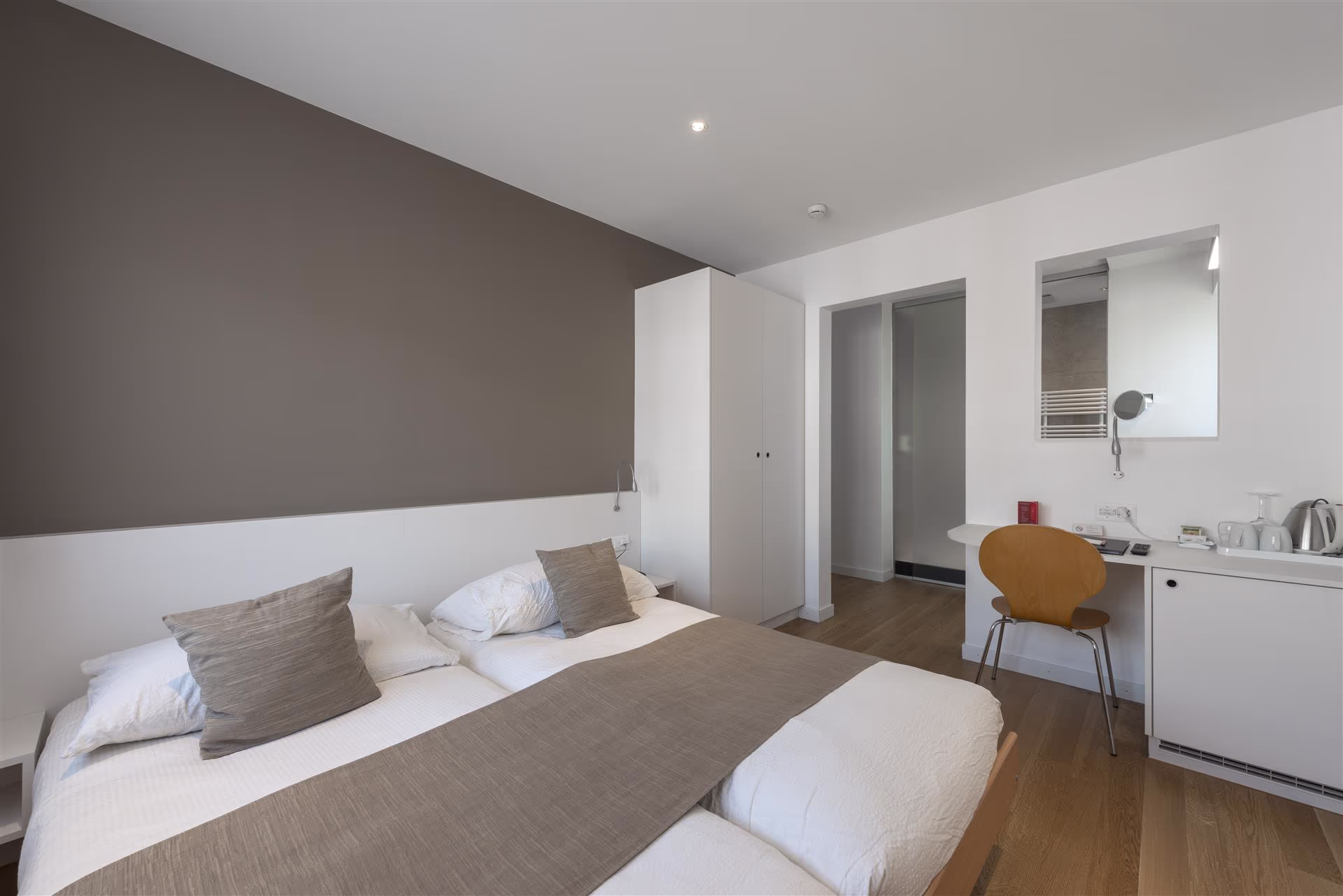 Chambre d’hôtel simple avec lit double, linge blanc et beige, armoire blanche, bureau avec chaise en bois, miroir et mini-cuisine à droite.