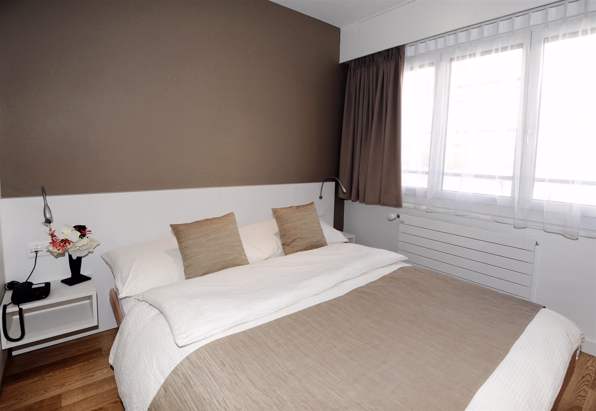 Chambre avec lit double, parure beige et blanche, deux coussins, table de chevet avec téléphone et vase, fenêtre avec rideaux bruns et voilages blancs.