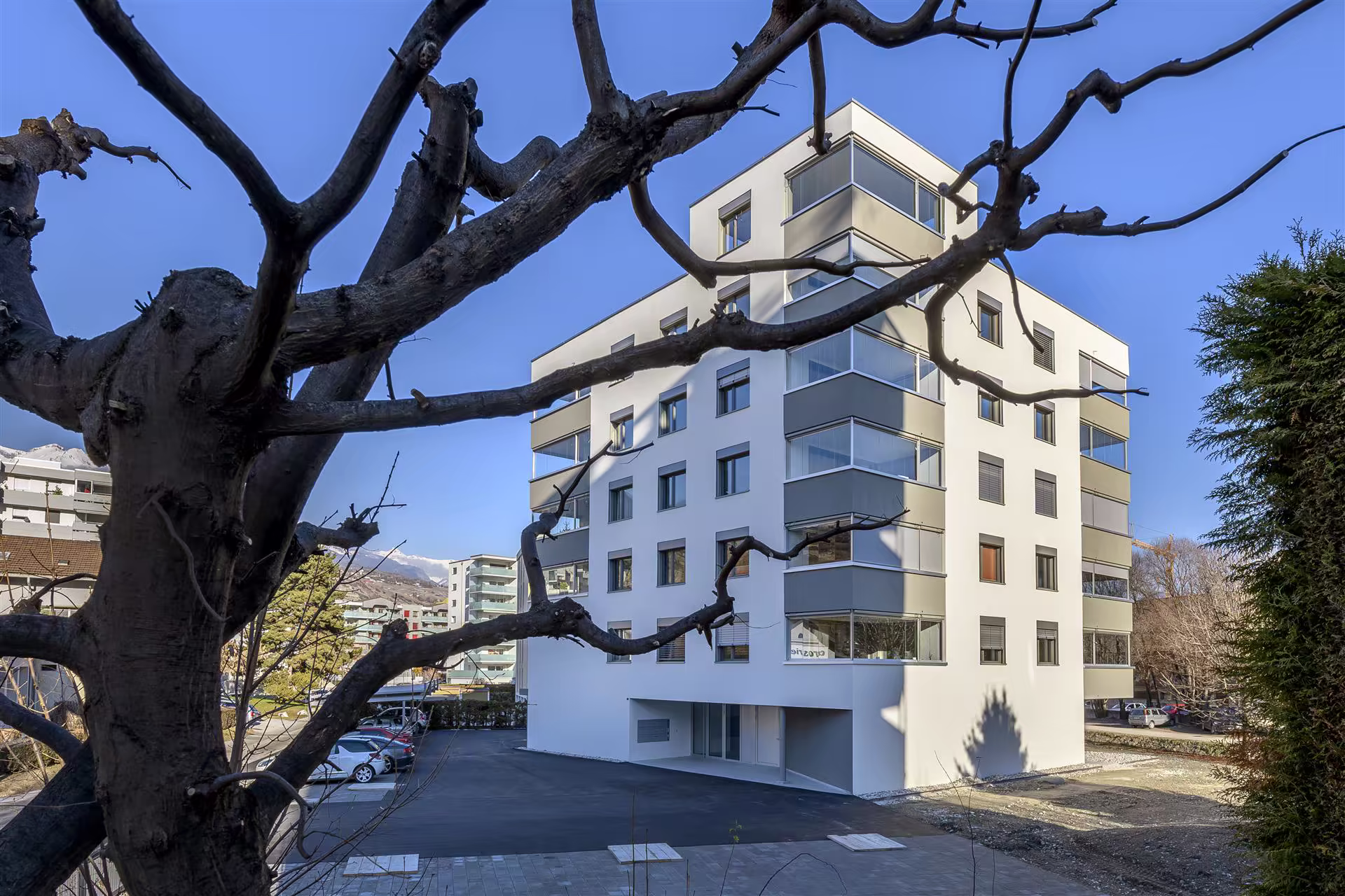 Bâtiment blanc moderne de cinq étages avec balcons vitrés, parkings à côté, arbres sans feuilles et ciel bleu clair.