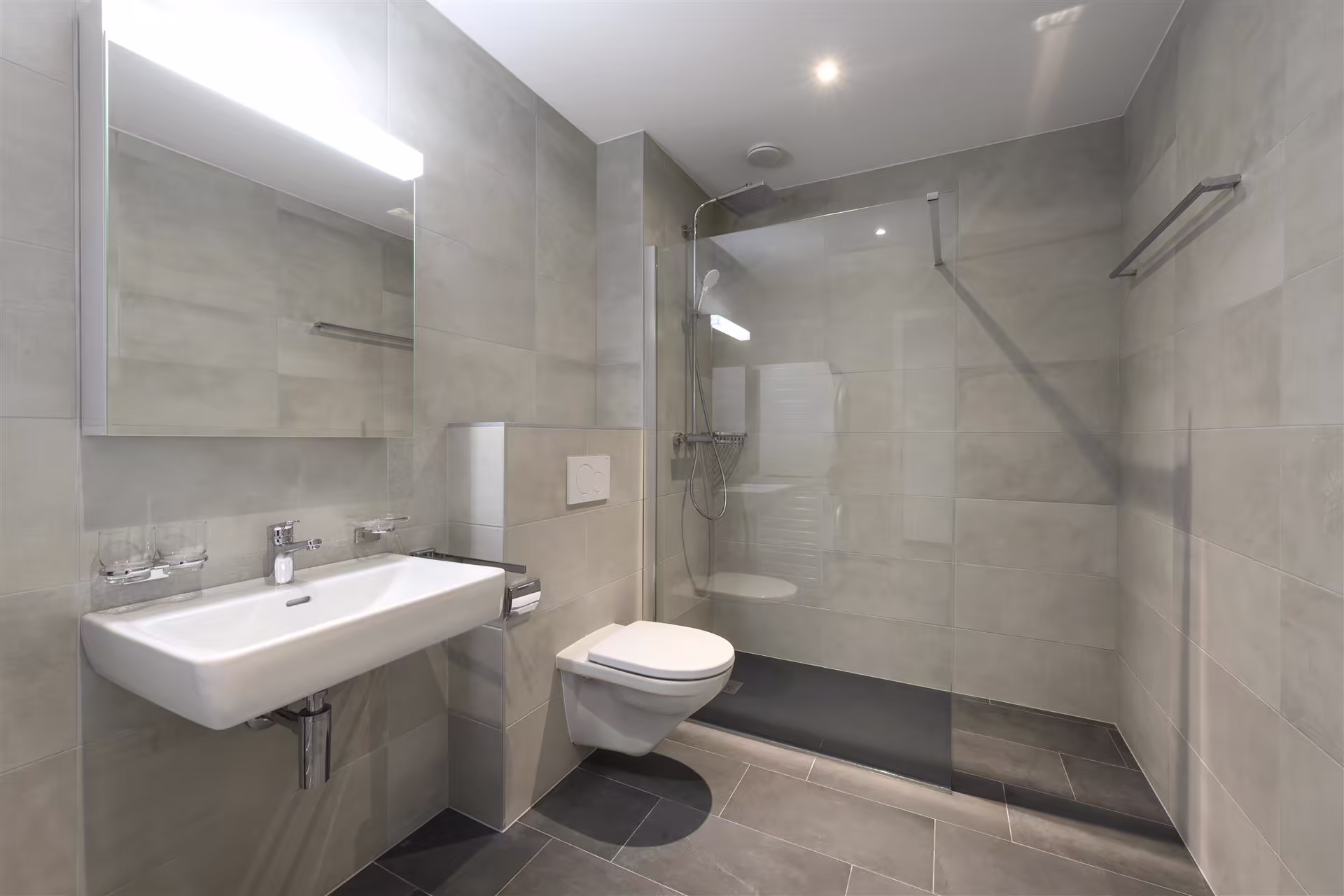 Salle de bain moderne avec toilettes suspendues, lavabo mural, grande douche à l'italienne avec paroi vitrée, carrelage gris clair et sol gris foncé.
