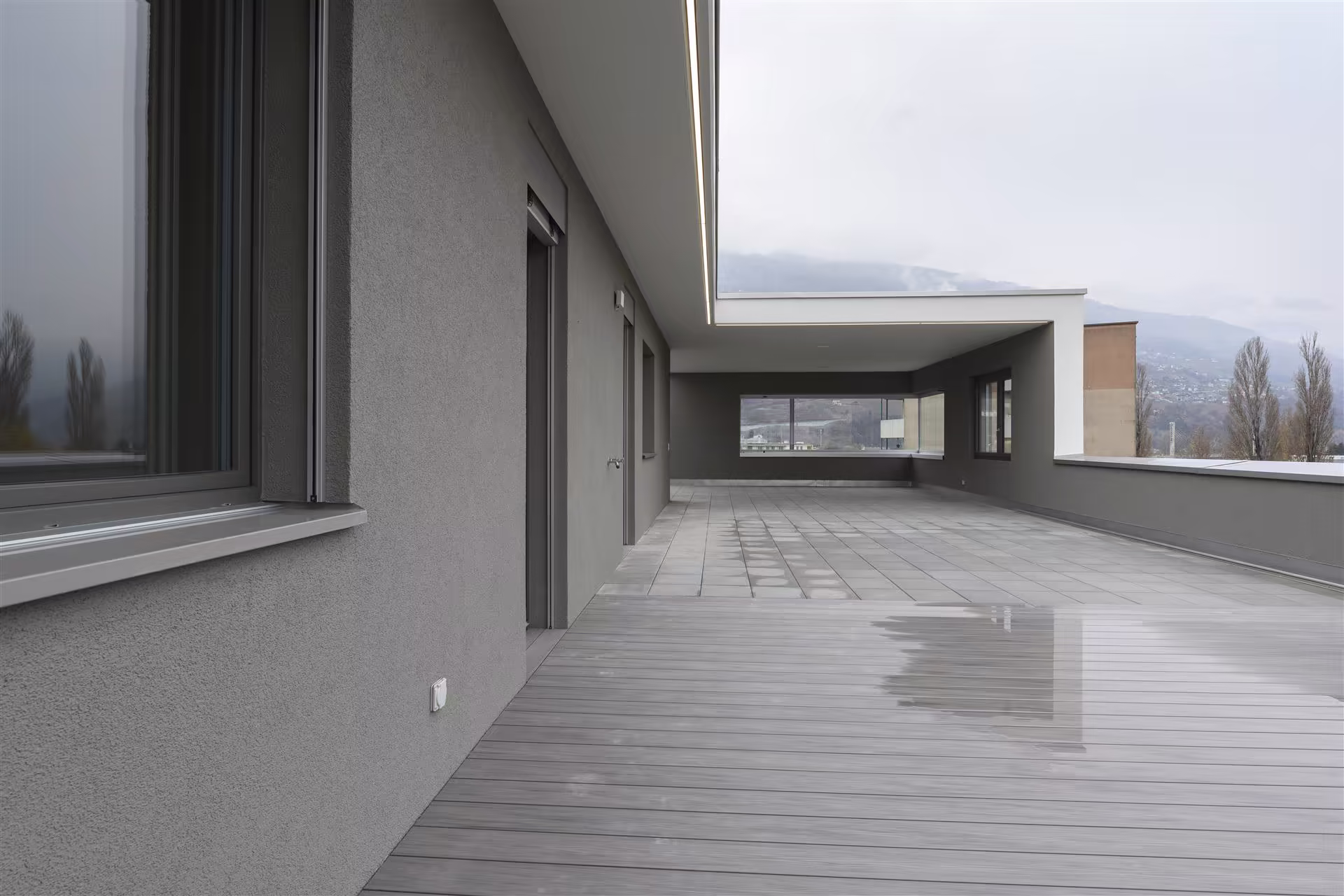 Grande terrasse moderne avec sol en lames de bois et dalles, murs gris, fenêtres, vue sur montagnes et ciel nuageux. Sol légèrement humide.