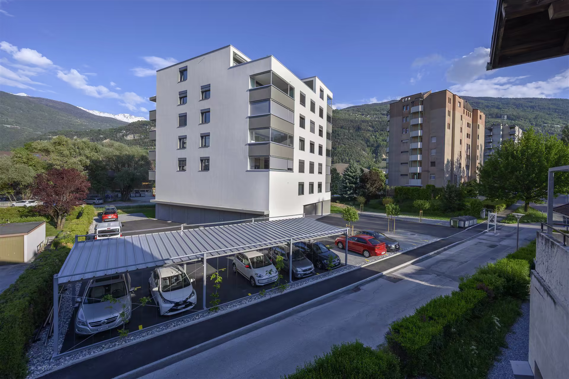 Résidence moderne à cinq étages avec parking abrité en avant, plusieurs voitures garées, montagnes en arrière-plan sous un ciel bleu partiellement nuageux.