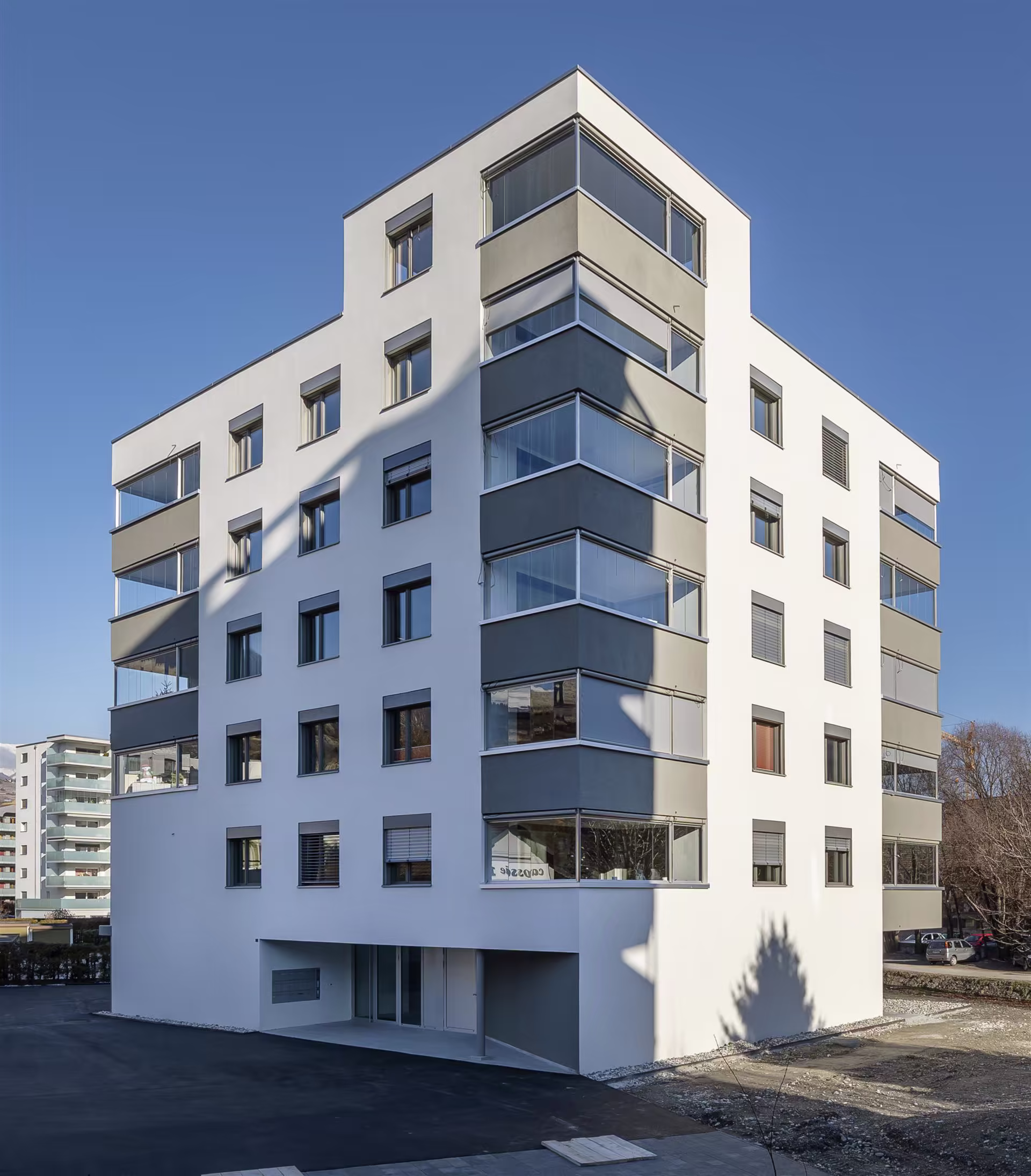 Bâtiment moderne à façades blanches et gris foncé, avec nombreuses fenêtres et balcons vitrés, sous ciel dégagé bleu.