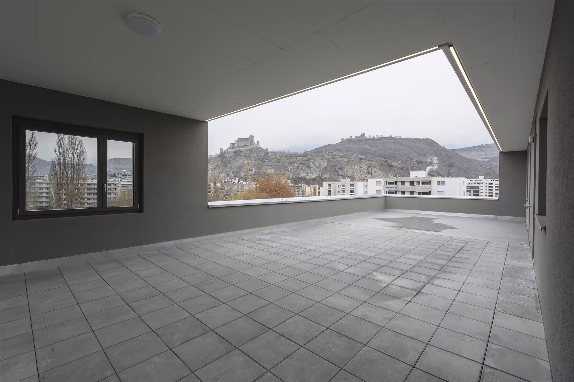 Grande terrasse couverte avec sol en carrelage gris, vue dégagée sur des collines rocheuses et des bâtiments résidentiels en arrière-plan.