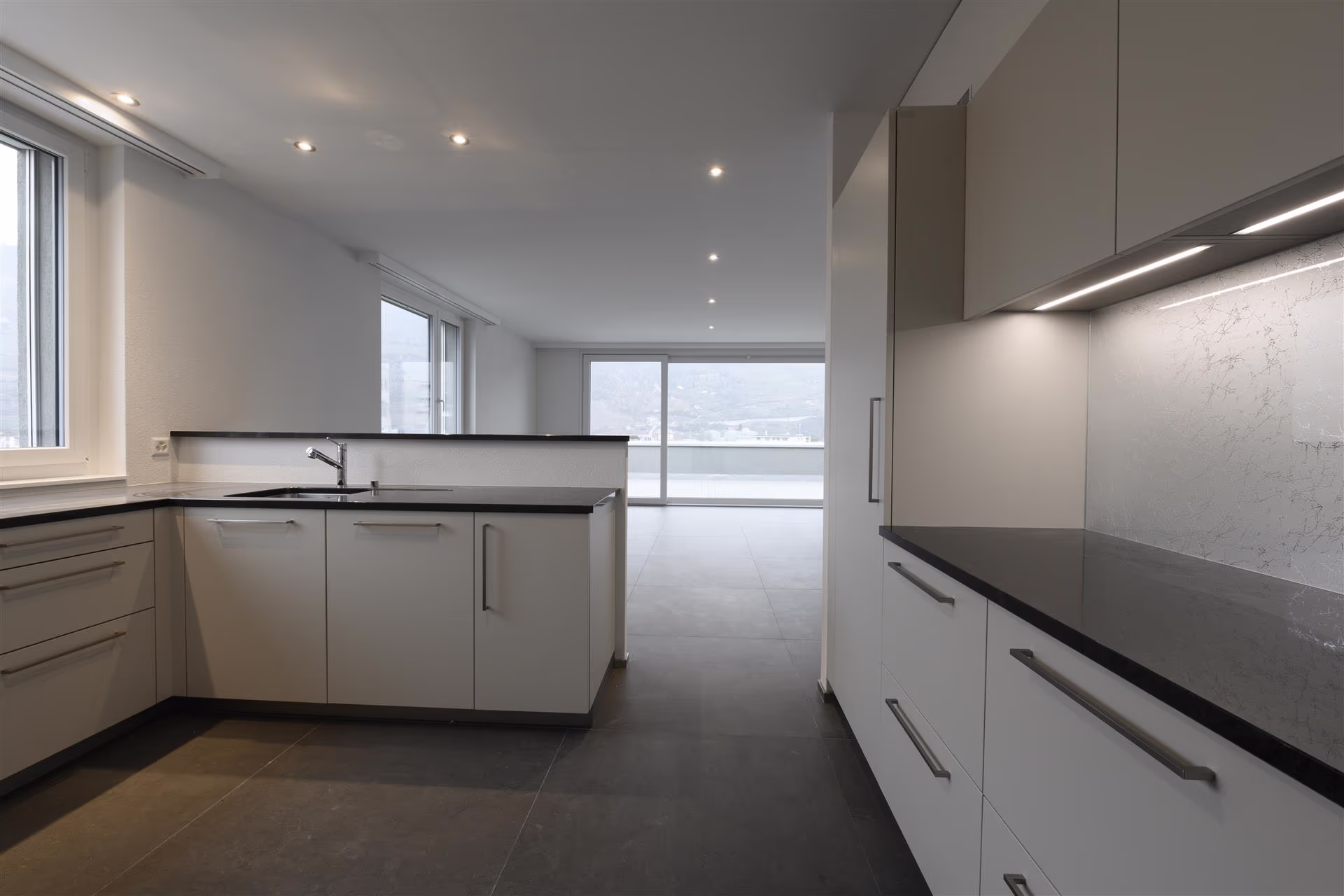 Cuisine moderne et minimaliste avec plans de travail noirs, armoires blanches, sol gris, fenêtres et baie vitrée donnant sur une terrasse et vue extérieure.