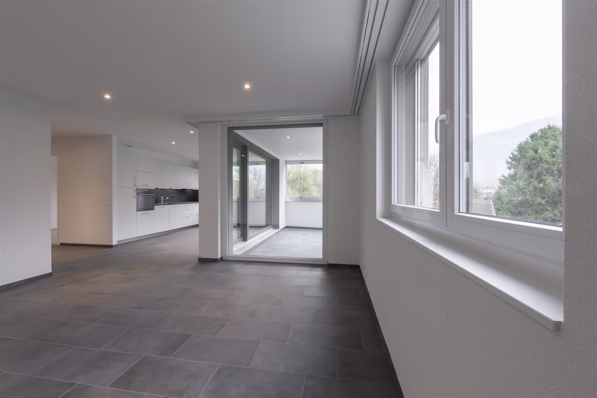Appartement vide avec sol en carrelage gris, cuisine blanche intégrée, grande baie vitrée et lumière naturelle venant d’une fenêtre à droite.