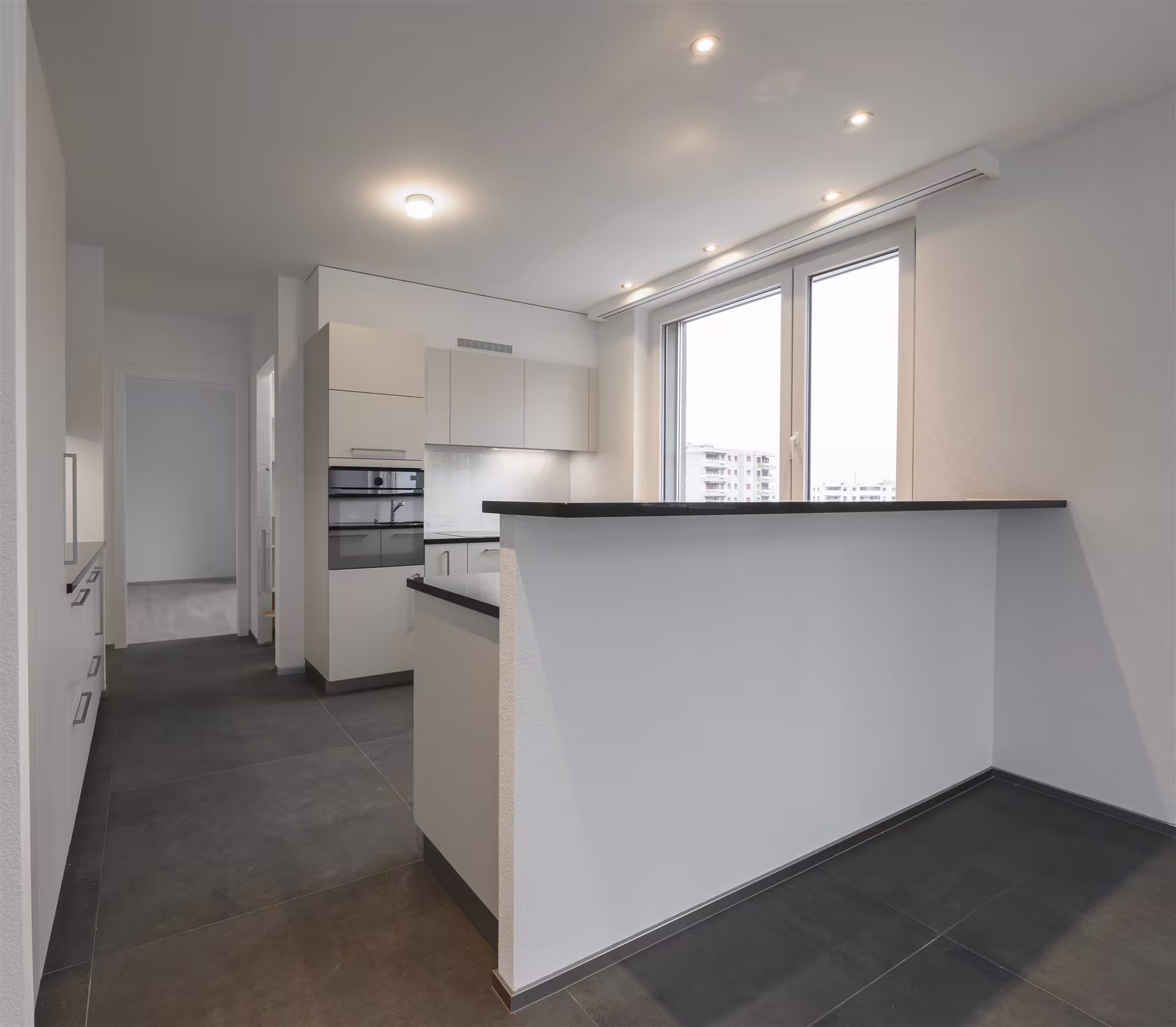 Cuisine moderne blanche avec plan de travail noir, carrelage gris foncé, fenêtre double vitrage et éclairage au plafond, sans meubles ni décoration.