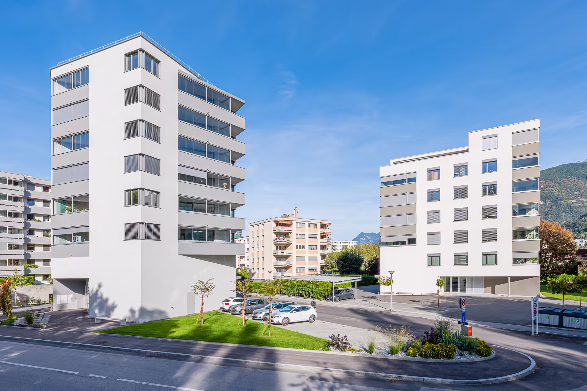Deux immeubles blancs modernes avec balcons vitrés, espace de stationnement avec voitures, arbres plantés et montagnes en arrière-plan sous un ciel bleu.
