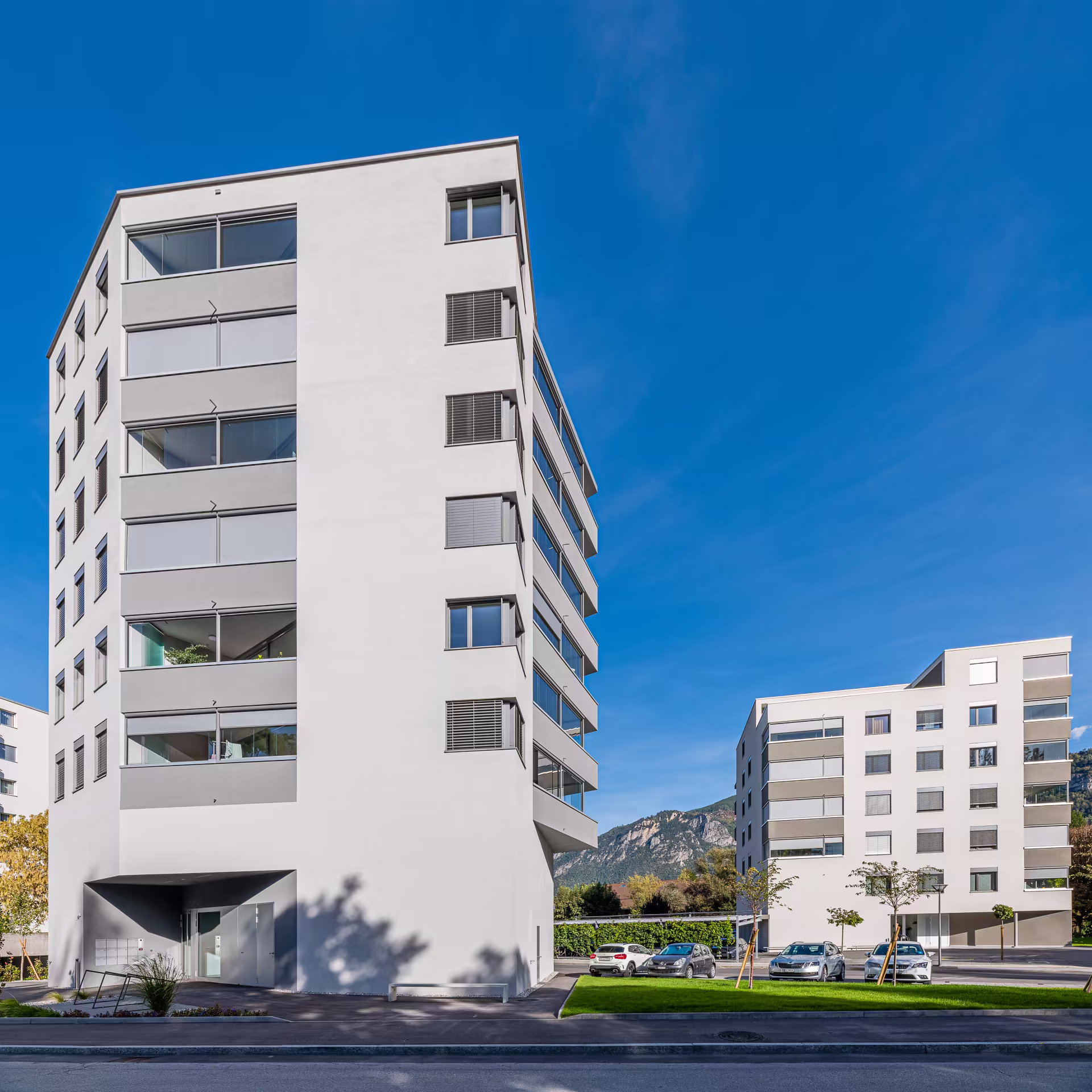 Vue en contre-plongée de deux immeubles modernes blancs avec balcons vitrés, voitures en stationnement, herbe verte et montagnes sous ciel bleu.