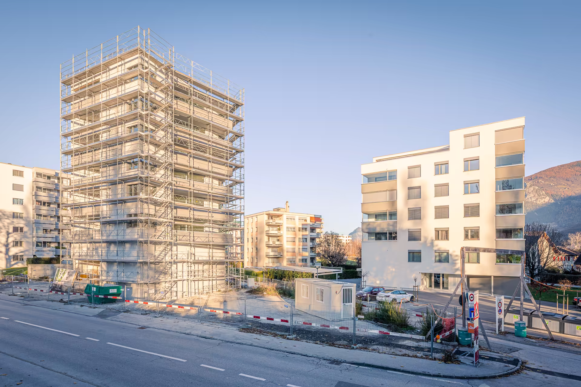 Immeuble en rénovation avec échafaudages à gauche, immeubles résidentiels à droite, route au premier plan et montagnes en arrière-plan.