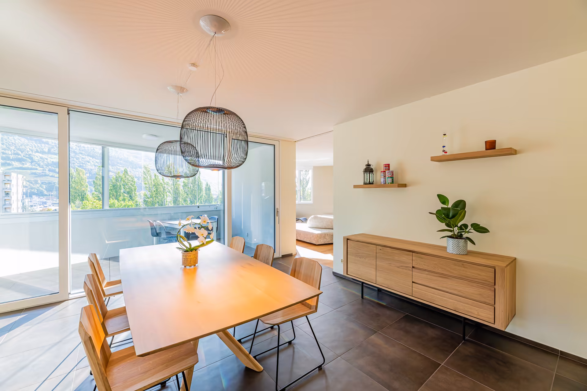 Salle à manger lumineuse avec grande table en bois, six chaises, deux suspensions noires, buffet bas et baie vitrée donnant sur une terrasse.