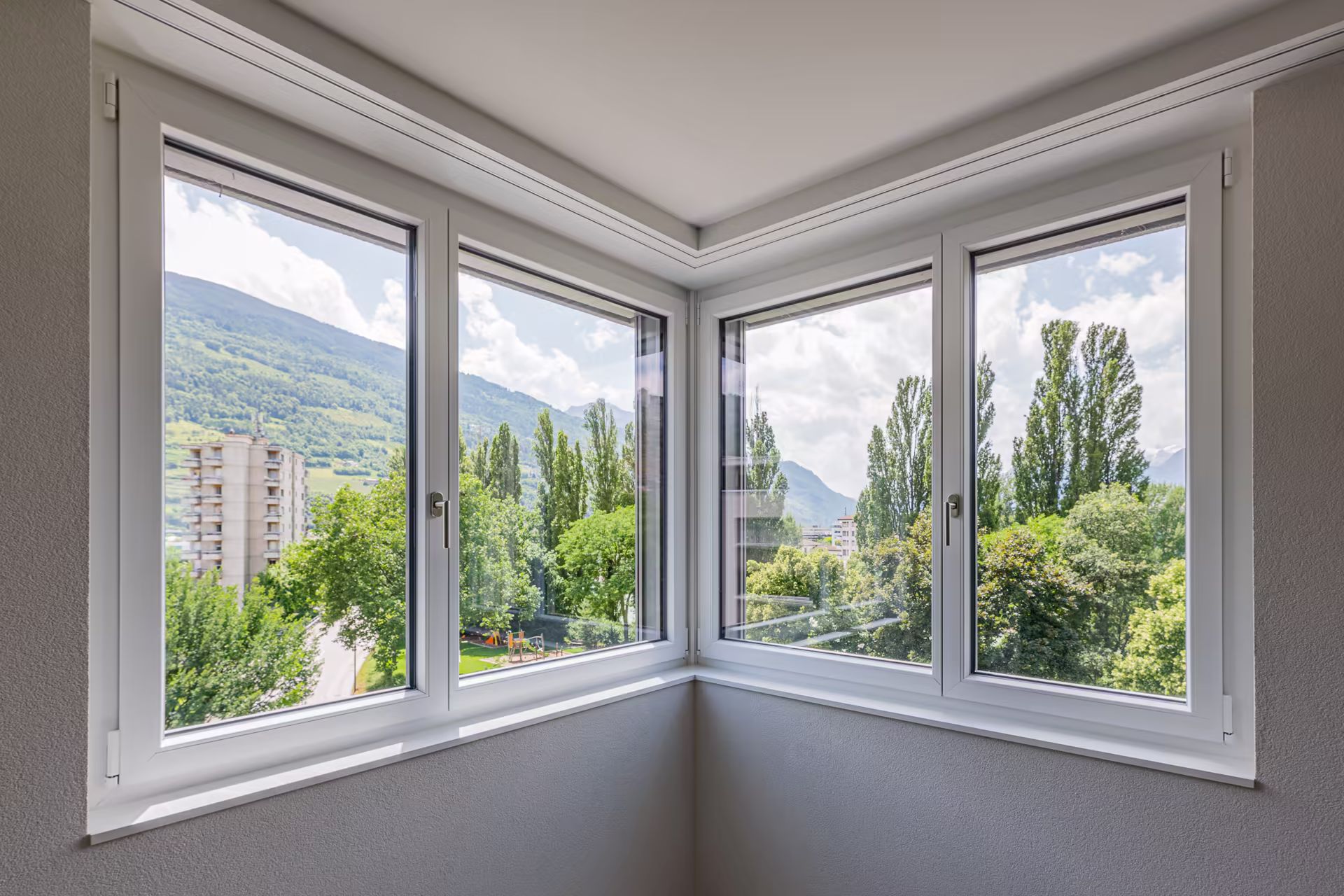 Fenêtre d’angle triple en PVC blanc donnant sur un paysage verdoyant avec arbres, immeuble, et montagne sous un ciel partiellement nuageux.