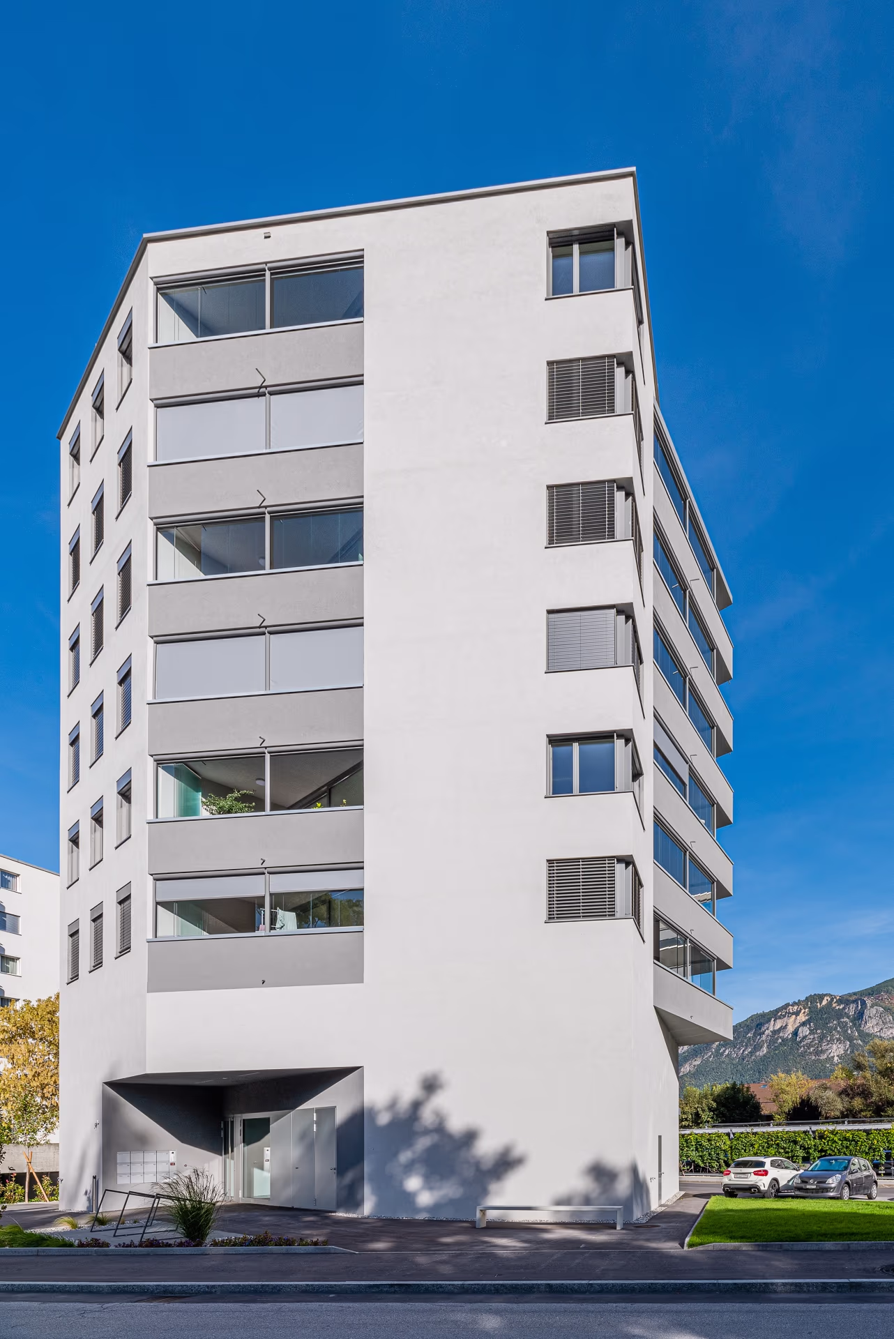 Bâtiment résidentiel moderne blanc à plusieurs étages avec balcons vitrés, entrée en retrait et ciel bleu, montagnes en arrière-plan.