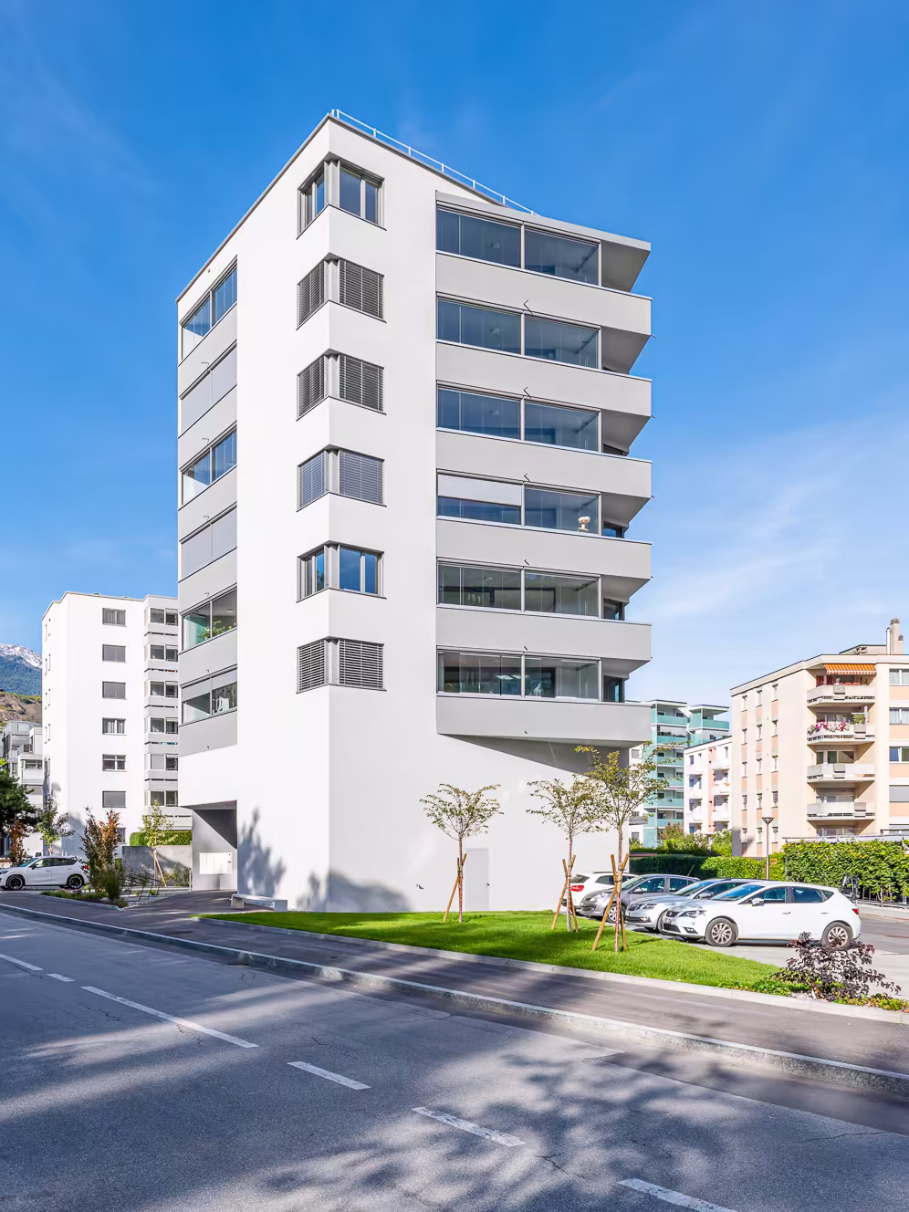 Bâtiment blanc moderne de six étages avec balcons vitrés, entouré de pelouses et de petites arbres, sous un ciel bleu sans nuages.