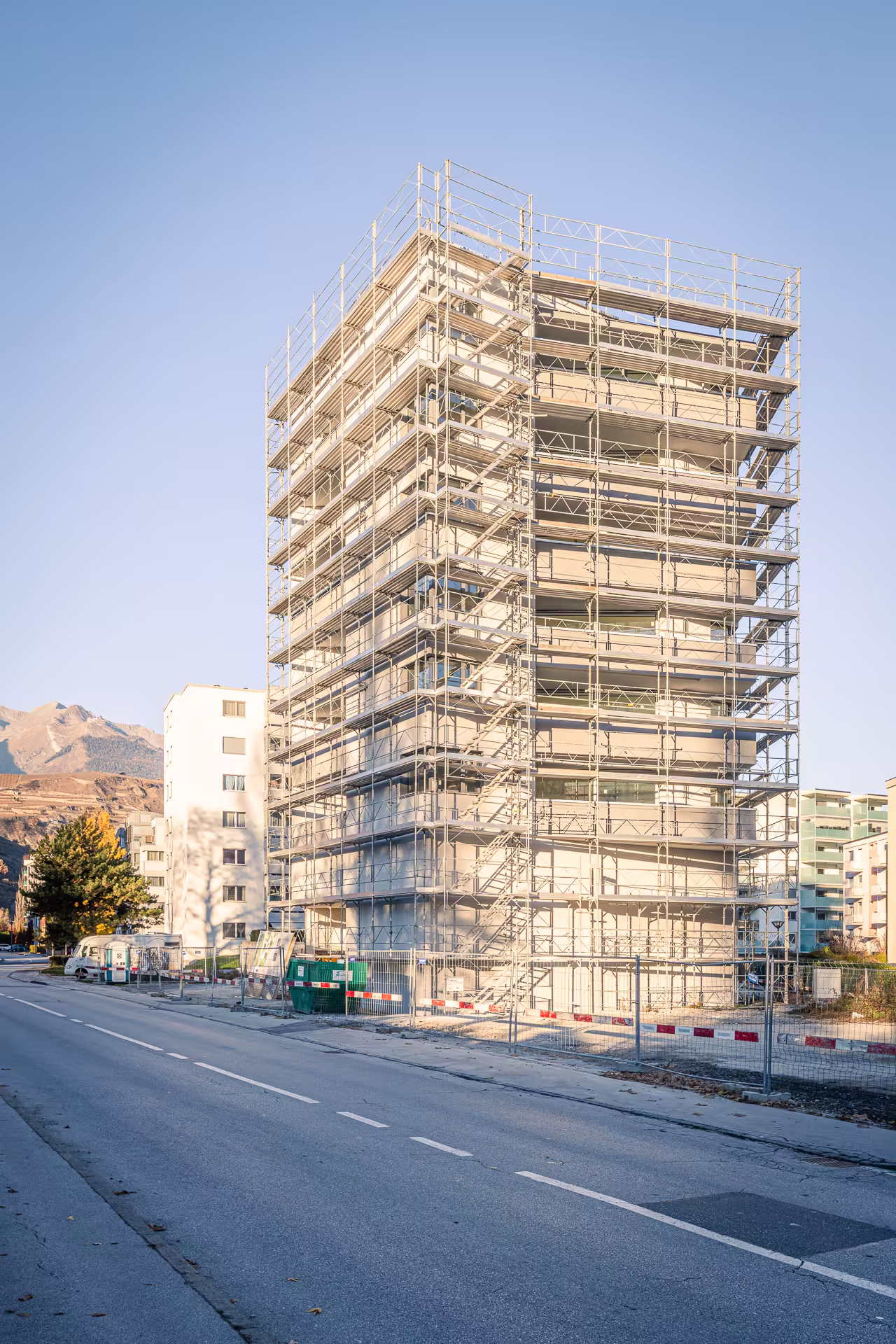 Immeuble entouré d’échafaudages en chantier sur un trottoir, avec une montagne en arrière-plan sous un ciel bleu clair.