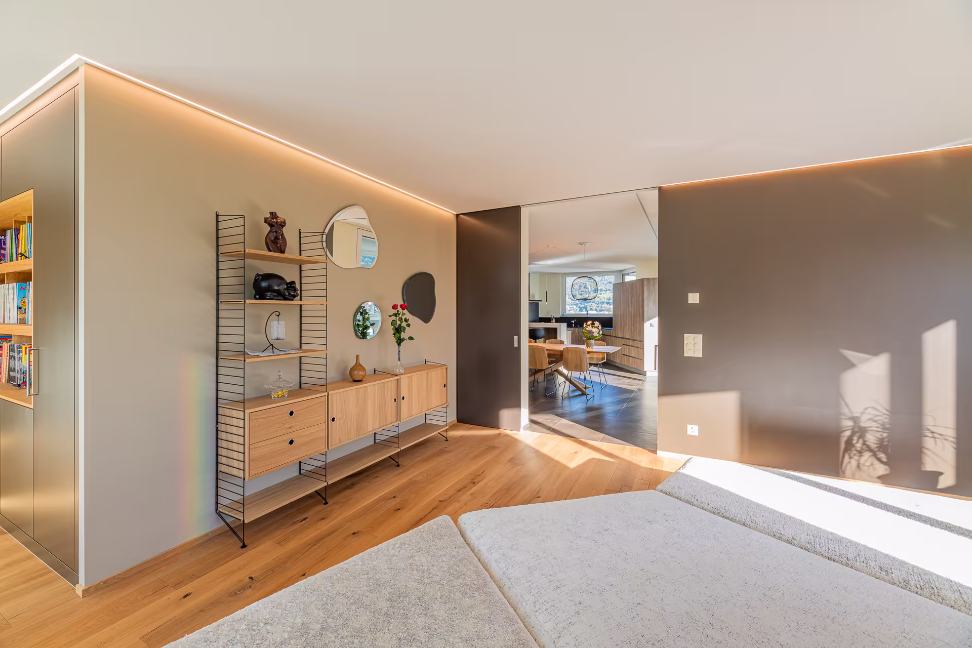 Chambre lumineuse avec parquet en bois, étagère murale avec décorations et miroir, ouverture vers une salle à manger moderne et lumineuse.