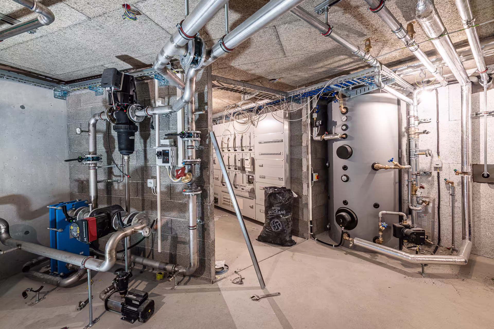 Local technique avec tuyauterie en acier, chaudière, pompe, panneau électrique et matériel de chauffage/installations industrielles.