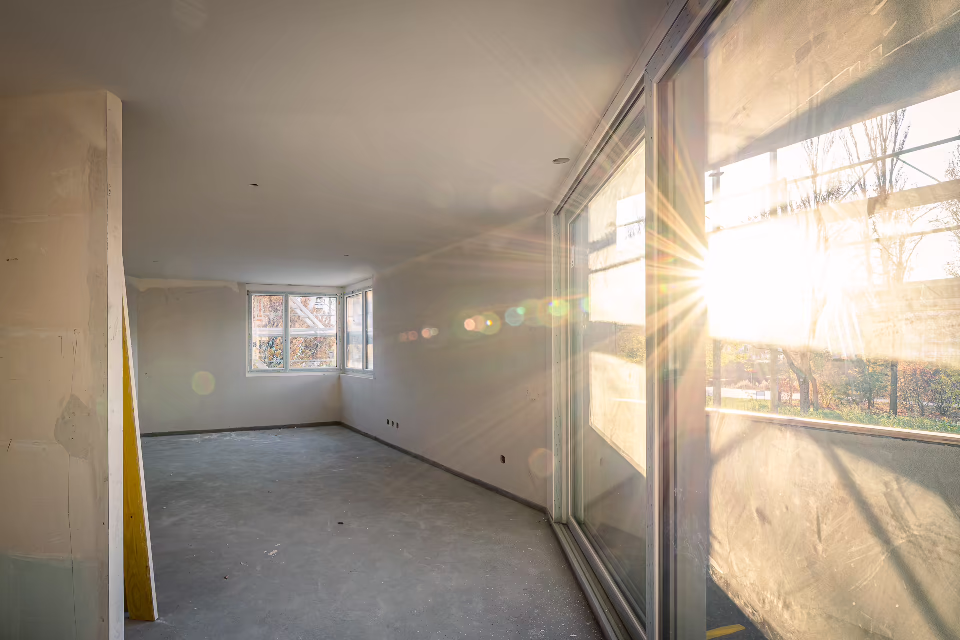 Intérieur d'une pièce en construction avec grandes fenêtres, sol en béton, murs non peints, et soleil pénétrant par une baie vitrée coulissante.