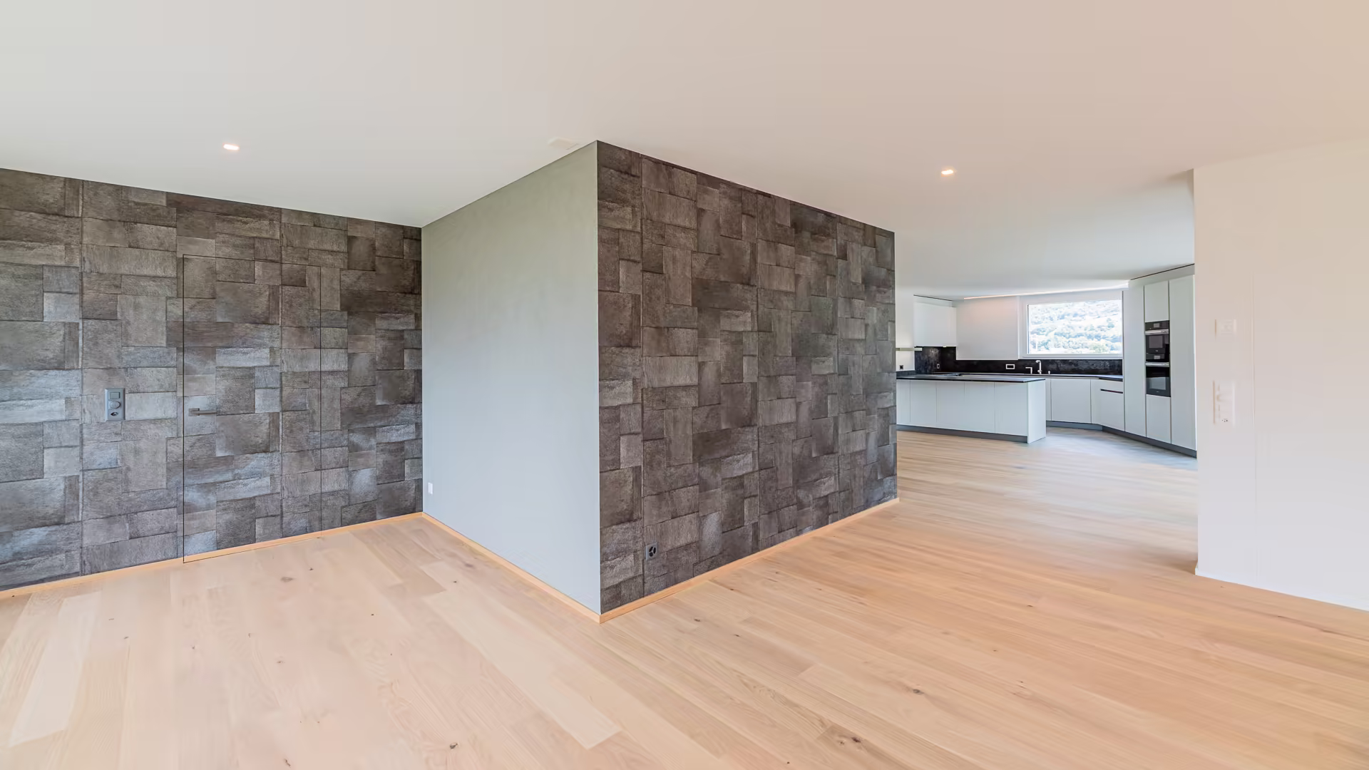 Coin d'une pièce moderne avec sol en parquet clair, mur en carrelage gris texturé, et cuisine blanche avec grand plan de travail et fenêtre.