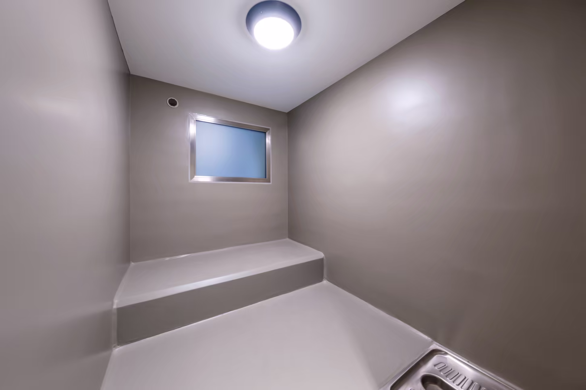 Petite cellule carcérale aux murs gris, avec une fenêtre opaque, une lumière au plafond, une assise en béton et un WC squat en acier inoxydable.