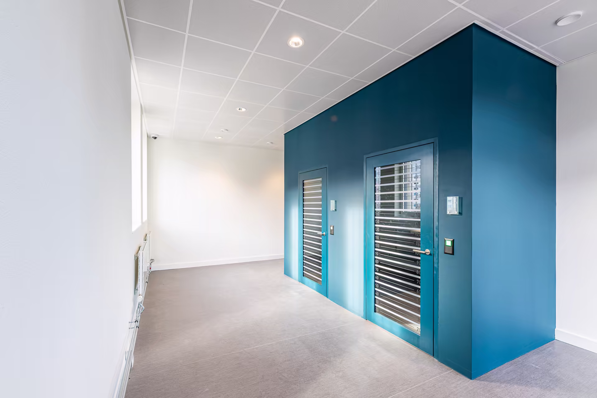 Couloir lumineux avec murs blancs et une structure bleue aux deux portes vitrées équipées de stores horizontaux.