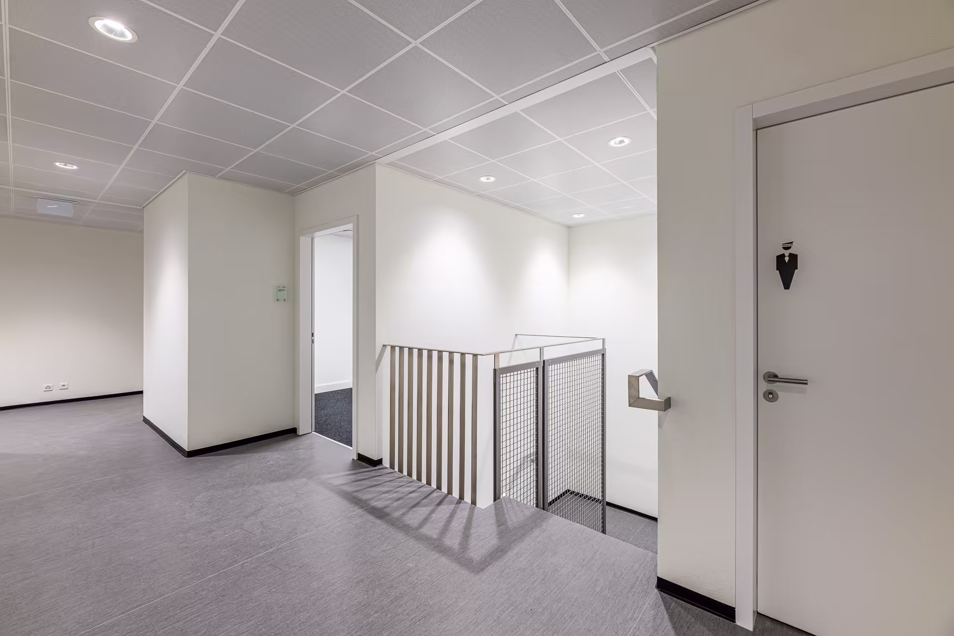 Couloir lumineux avec sols gris, murs blancs, porte de toilettes hommes à droite et escalier central protégé par une rampe métallique.