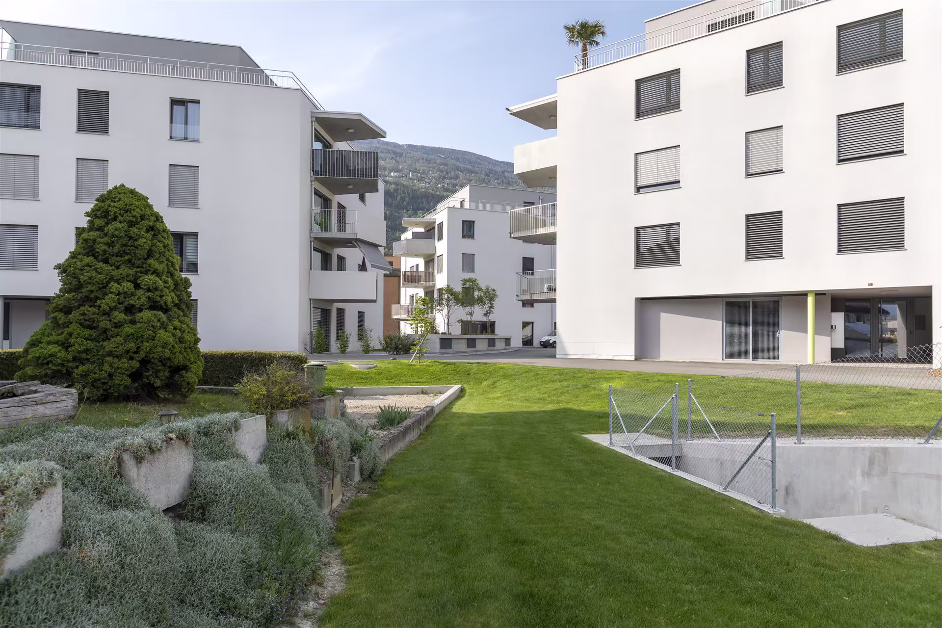 Cour intérieure d’un quartier résidentiel moderne avec bâtiments blancs, pelouse verte, arbustes et montagnes en arrière-plan.