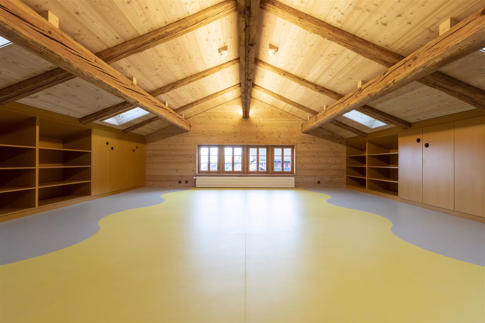 Grenier aménagé avec sol jaune et gris, murs et plafond en bois clair, grande fenêtre et rangements intégrés sous pente avec éclairage naturel.