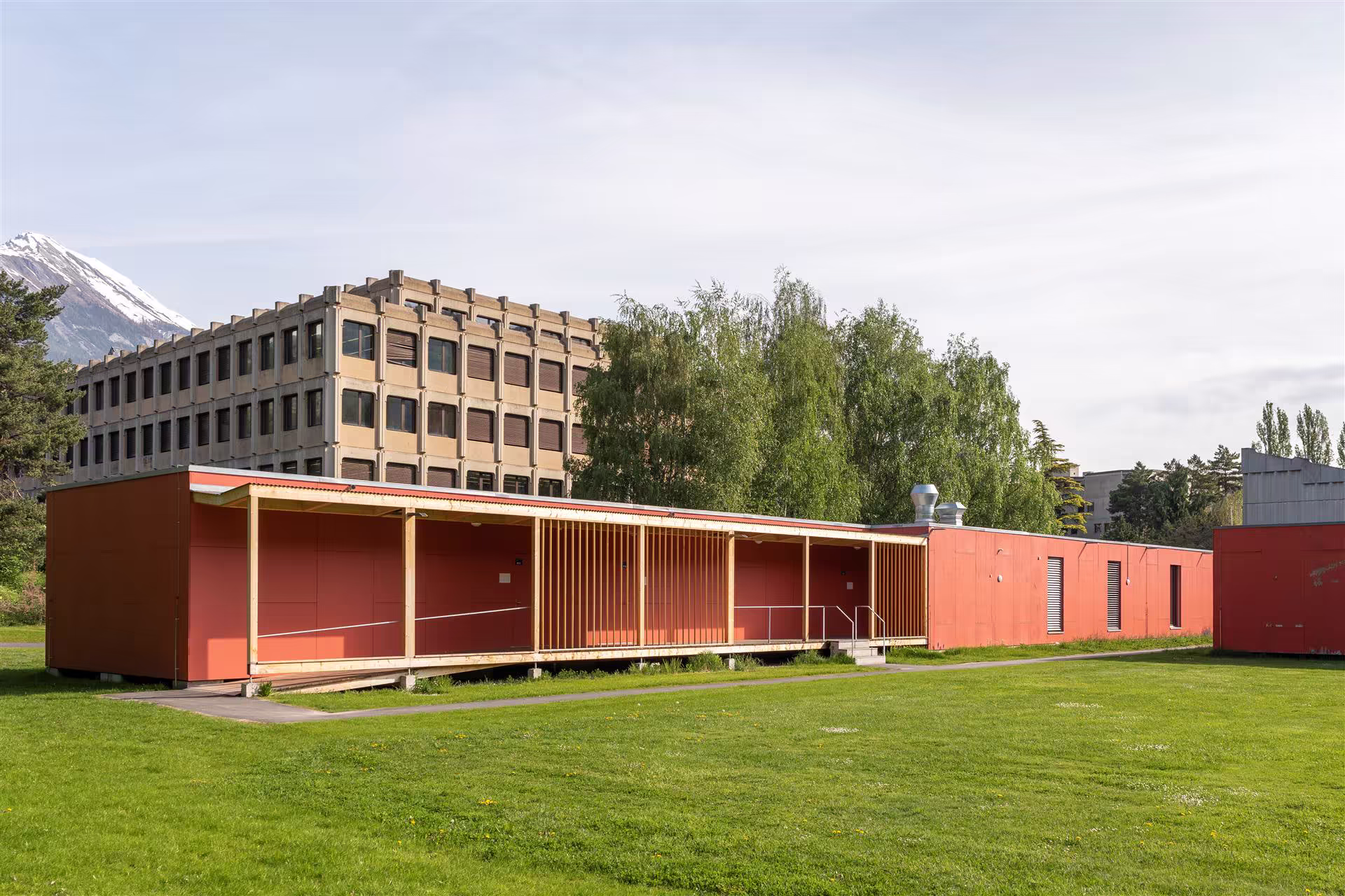 Bâtiment long rouge avec une galerie en bois, pelouse verte au premier plan, arbres et immeuble cubique en béton à l’arrière-plan, montagne enneigée au loin.