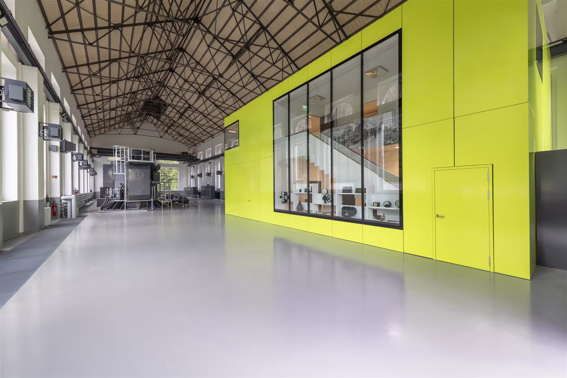 Grand espace industriel désaffecté avec un plafond métallique et une structure jaune vif contenant une pièce vitrée avec escalier et objets d'exposition.