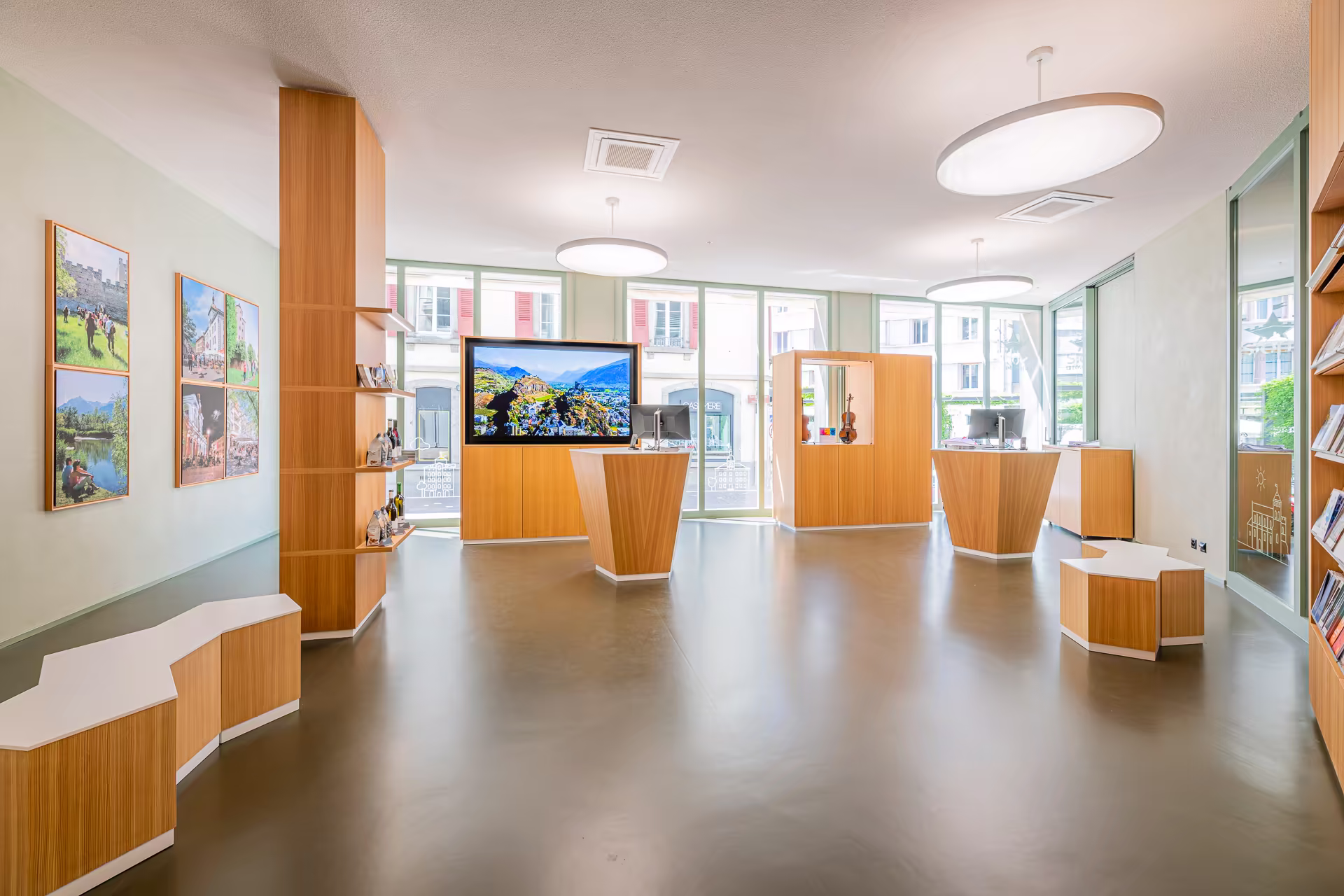 Espace lumineux avec comptoirs en bois, bancs modulables, vitrine avec violon, grand écran affichant un paysage montagneux, murs ornés de photos.