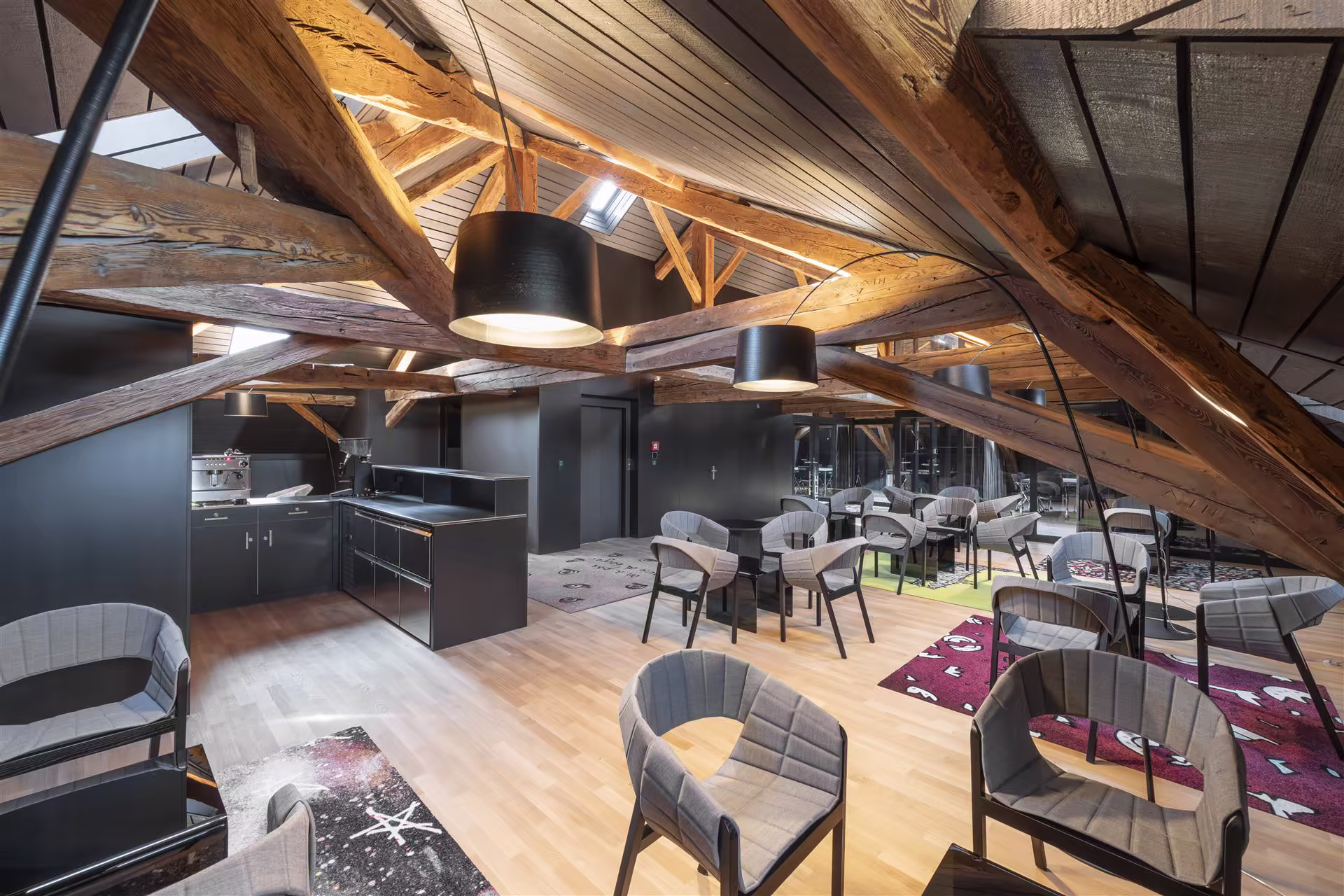 Espace lounge sous des poutres en bois apparentes avec des fauteuils gris, tables noires, lampes suspendues noires et moquettes colorées.