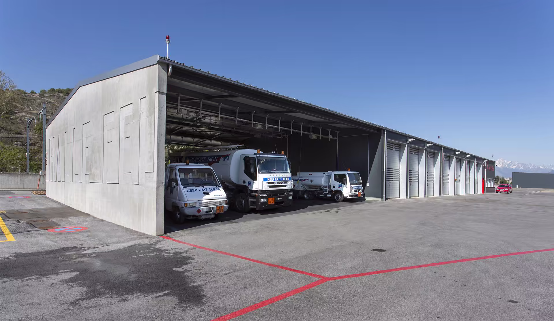 Dépôt extérieur avec trois camions-citernes blancs garés sous un grand abri métallique, sol en béton et ciel bleu dégagé.