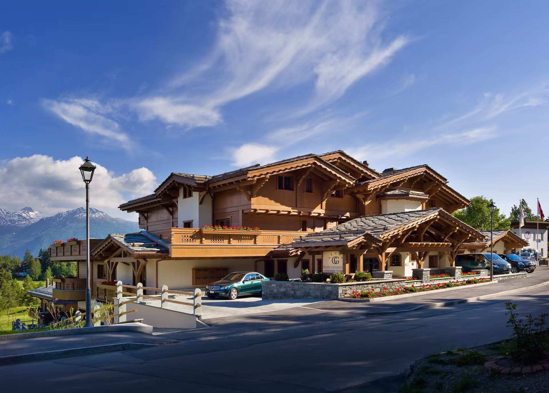 Grand chalet en bois clair avec balcon fleuri, voitures stationnées, lampadaires, montagne enneigée en arrière-plan sous un ciel bleu avec nuages légers.