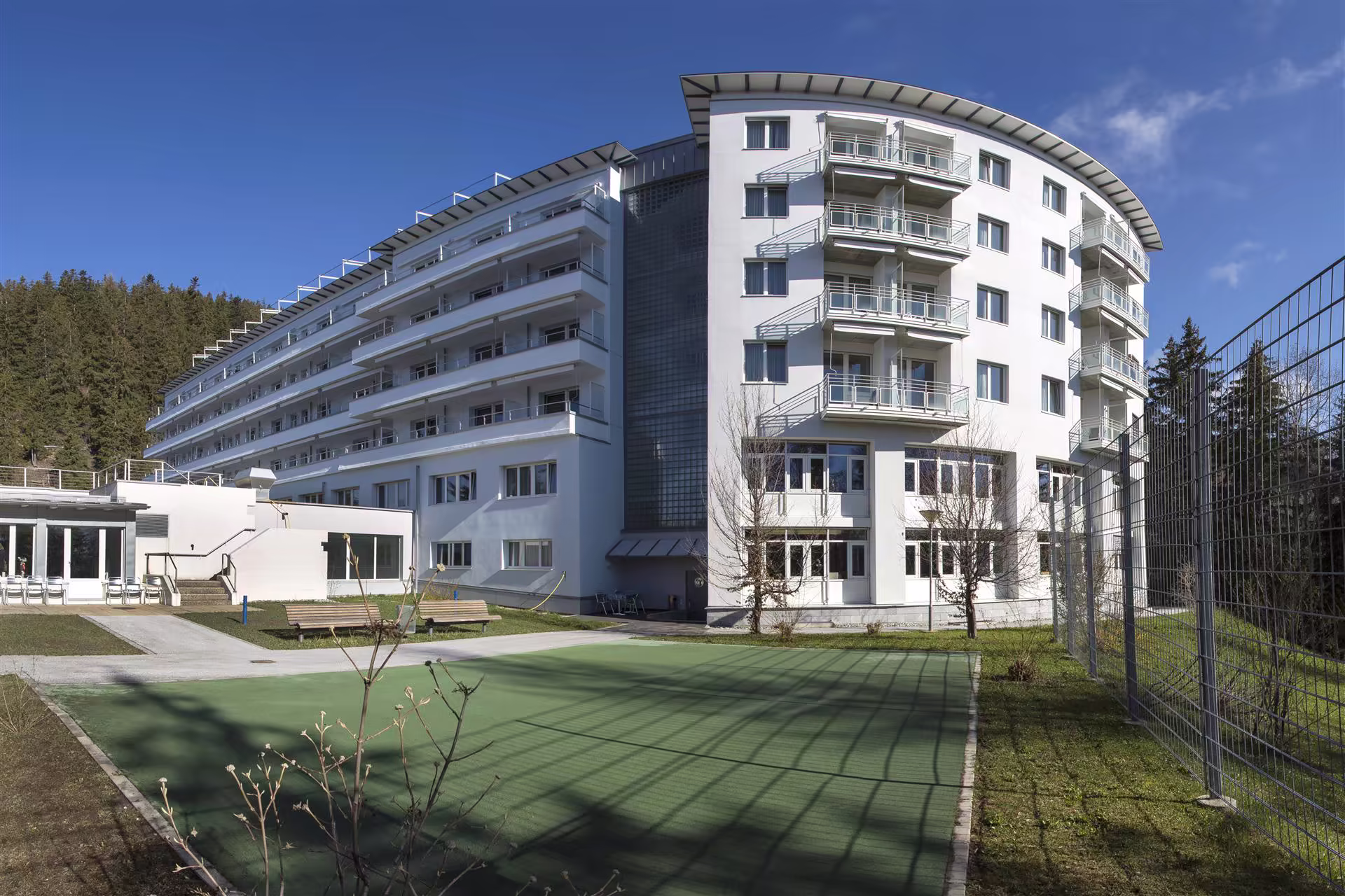 Bâtiment blanc moderne de plusieurs étages avec balcons et fenêtres, entouré d’un espace vert et d’une forêt sous un ciel bleu clair.