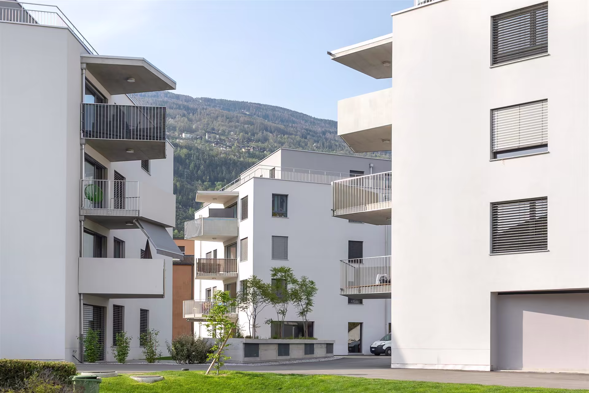 Complexe résidentiel moderne avec façades blanches, balcons métalliques, pelouse verte et collines boisées en arrière-plan sous un ciel clair.