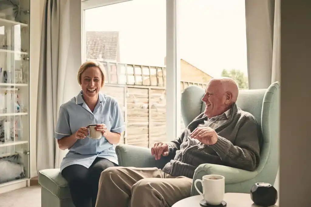 A carer and an older man laugh together over cups of tea at home