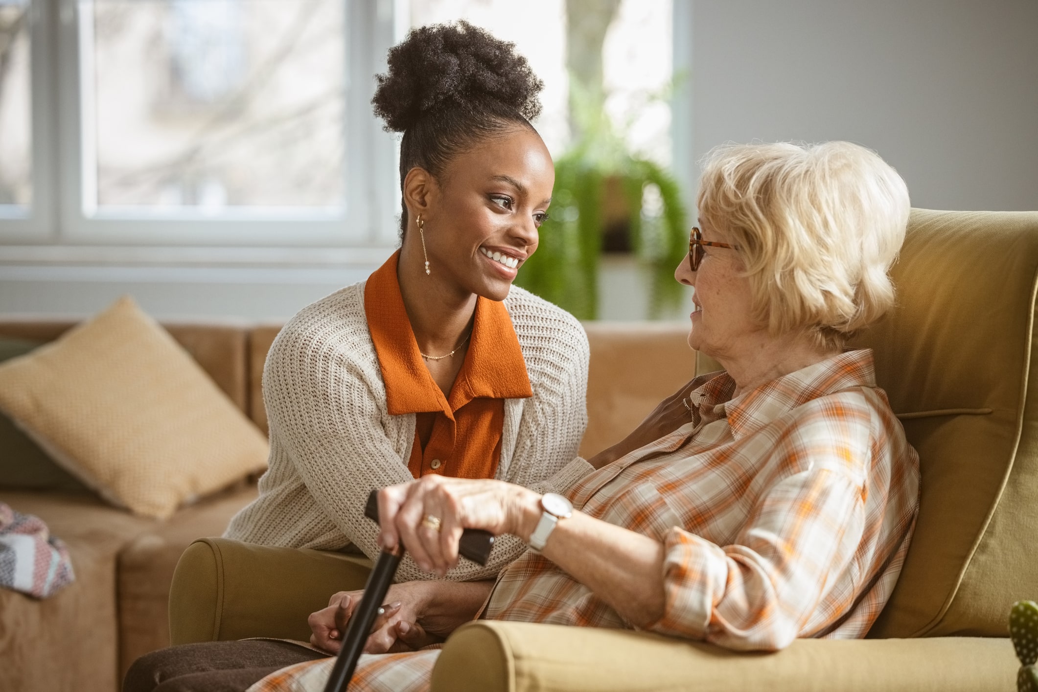 A smiling carer leans in to talk with an older woman in an armchair