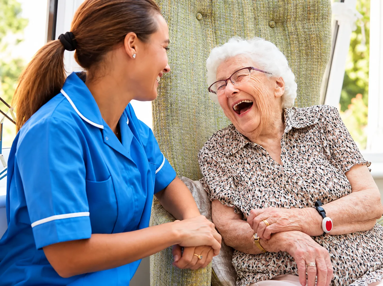 A carer in a blue uniform laughs with an older woman sitting in an armchair