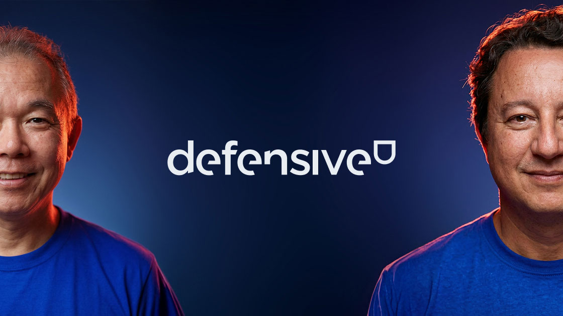 Dois homens sorrindo com camisetas azuis e logo defensivo no centro em fundo azul escuro.