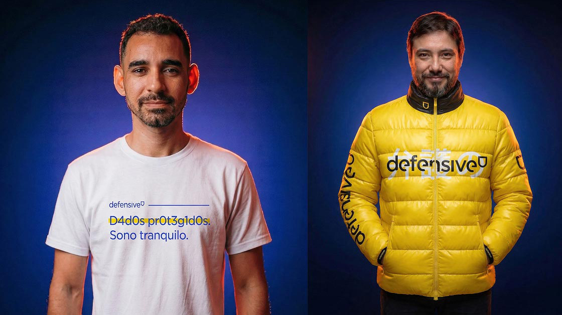 Dois homens sorrindo com camiseta branca e jaqueta amarela com texto 'defensive' em fundo azul.