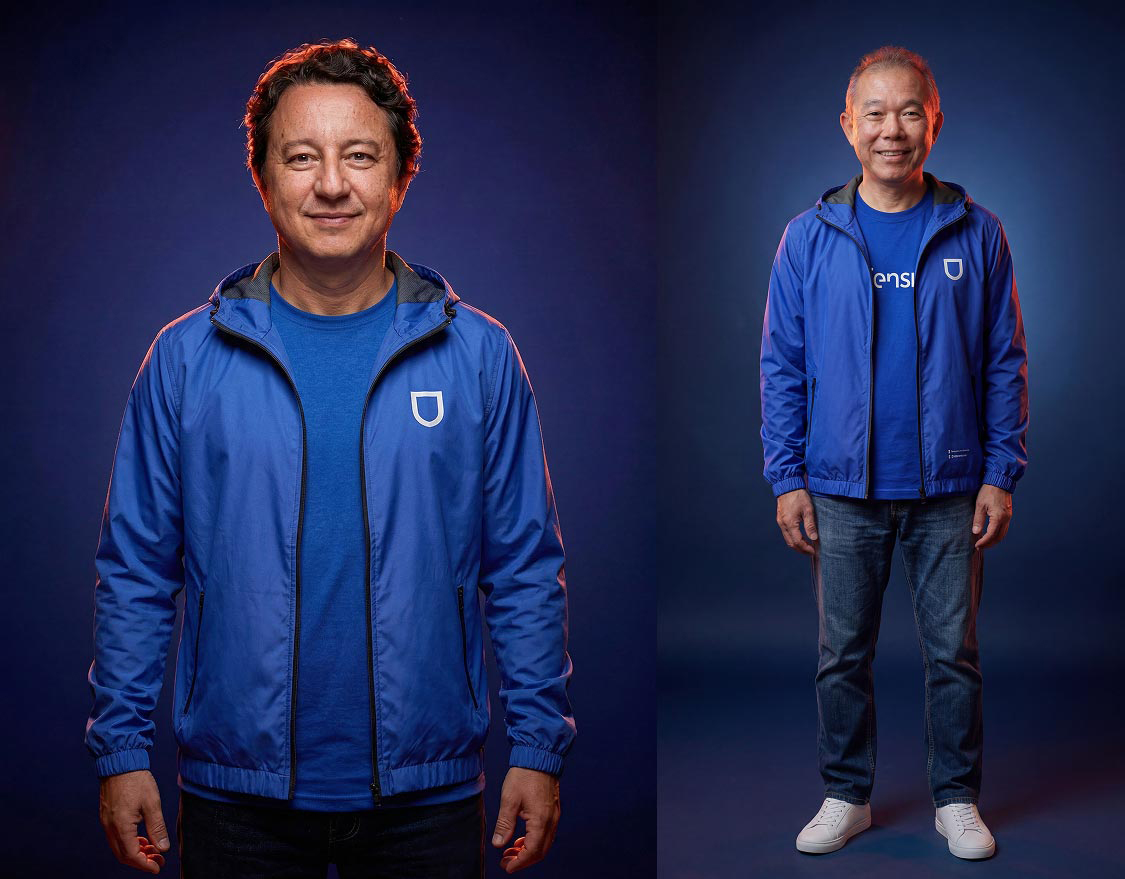 Dois homens sorrindo, vestindo jaquetas azuis com capuz e camiseta azul, em fundo azul escuro.