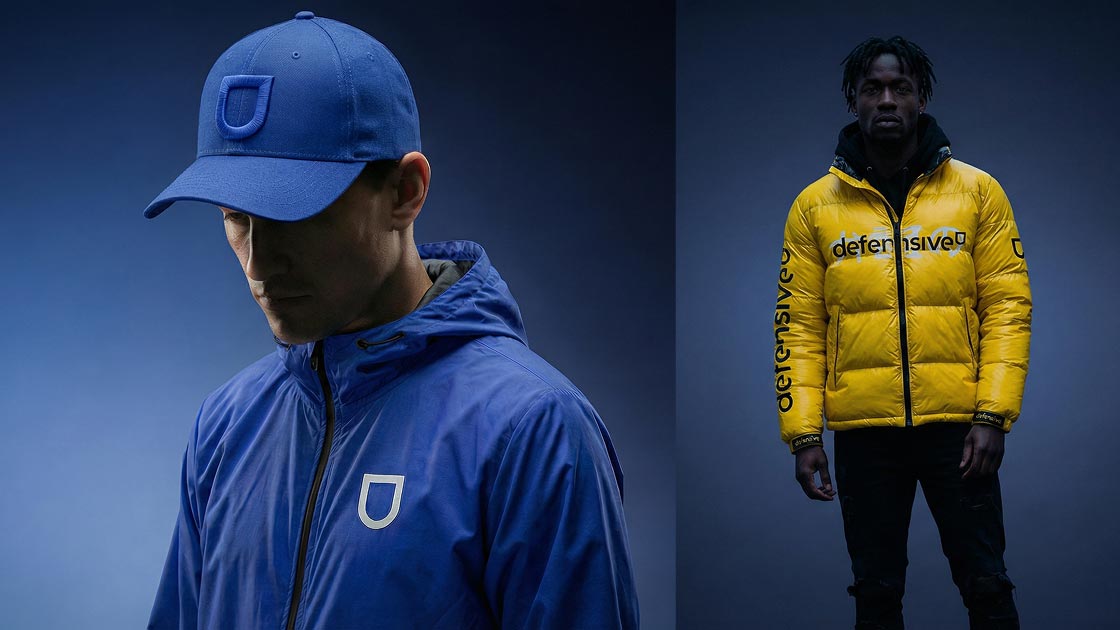 aplicação do signo defensive em um bone azul com linha texturada azul e jaqueta corta-vento azul com signo branco. aplicação da marca defensive centralizada no peito em um jaqueta puffer amarela.