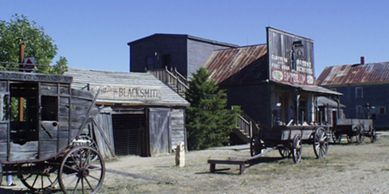 Old western town scene with wooden buildings, wagons, and dry grass under blue sky.