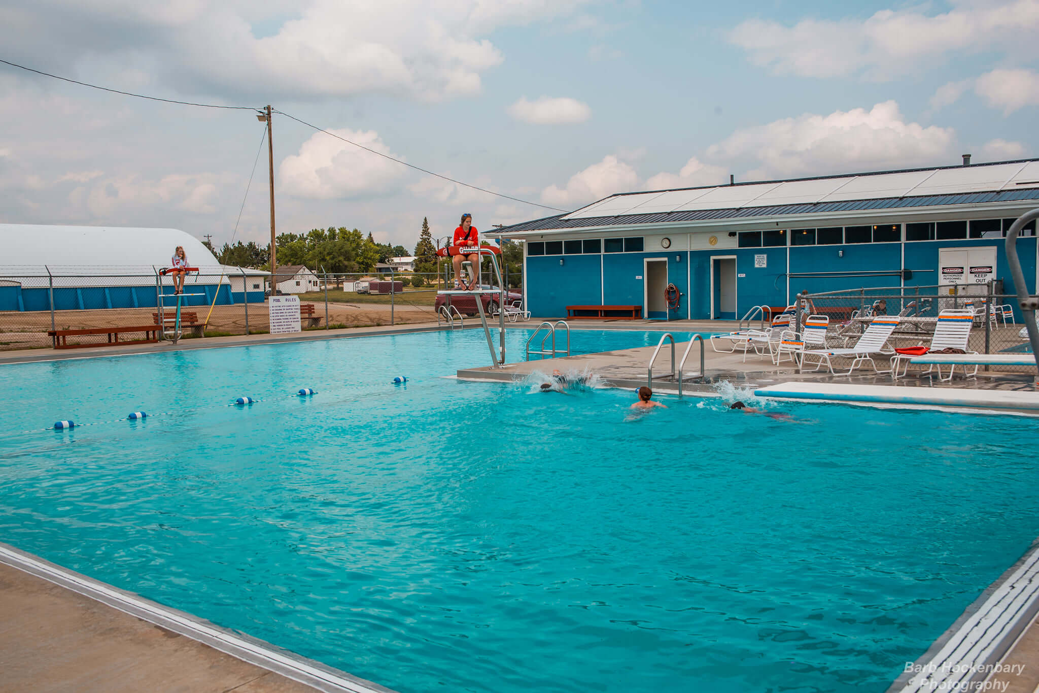 Two lifeguards watch over an outdoor pool where three people swim near the poolside.
