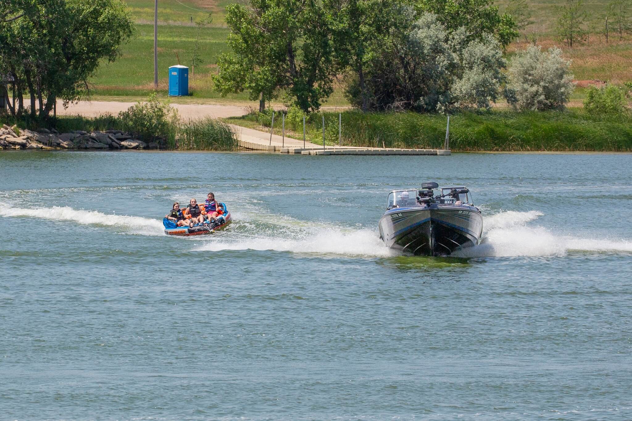 Boat pulling an inflatable tube with four people riding on a lake near a dock.