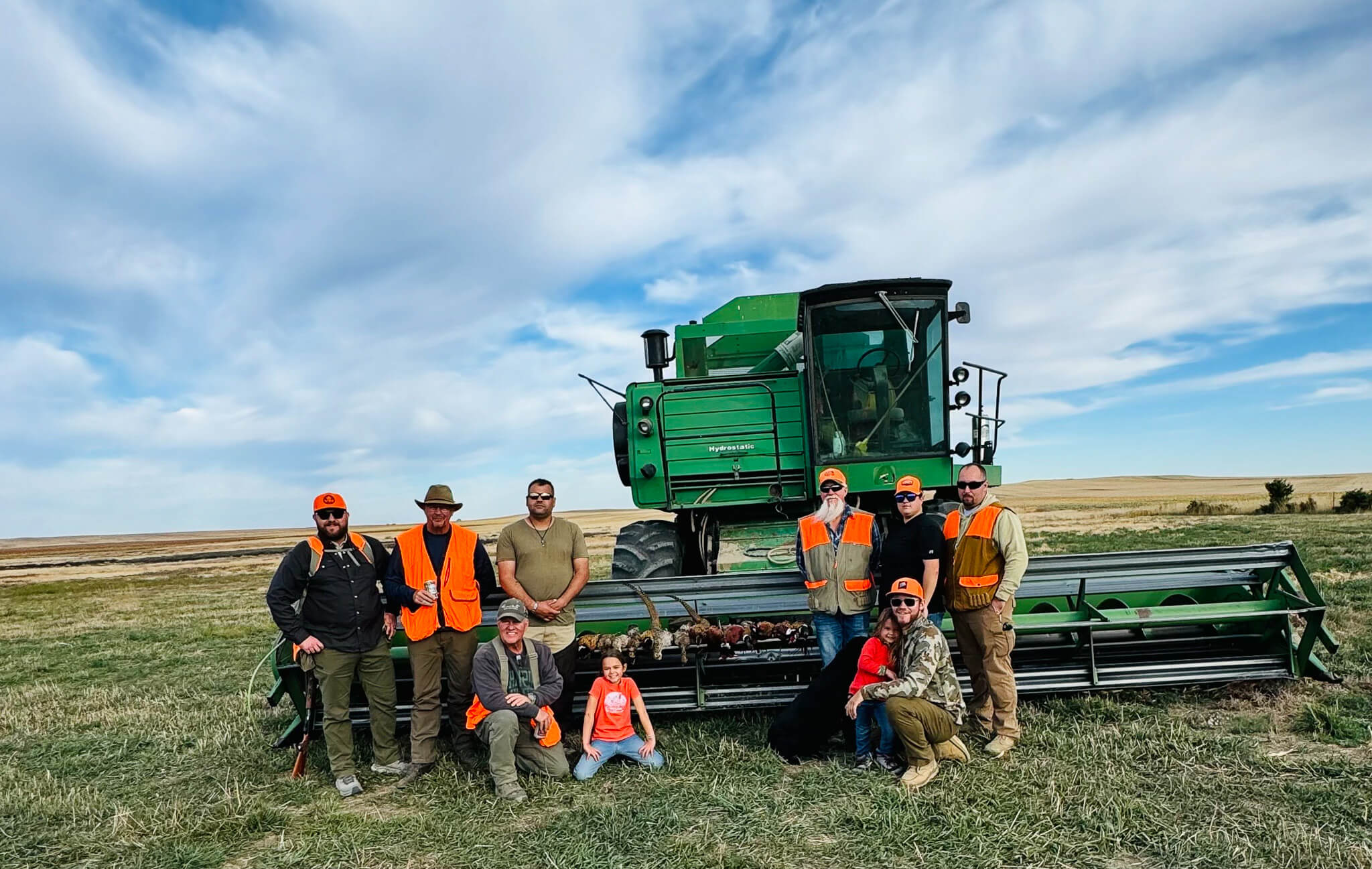 Group of people posing in front of green agricultural machinery on grassy field under blue sky.