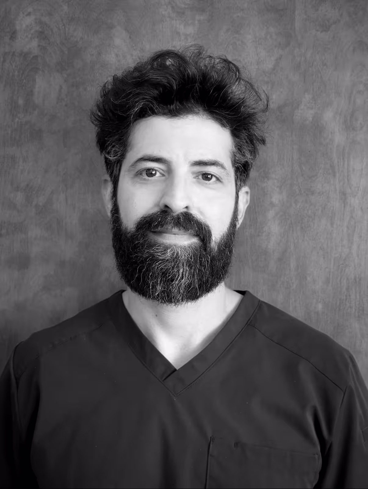 Black and white portrait of a man with a full beard and curly hair wearing a dark V-neck shirt.